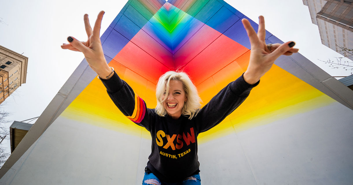 girl in an sxsw sweatshirt holding up peace signs in front of a rainbow background
