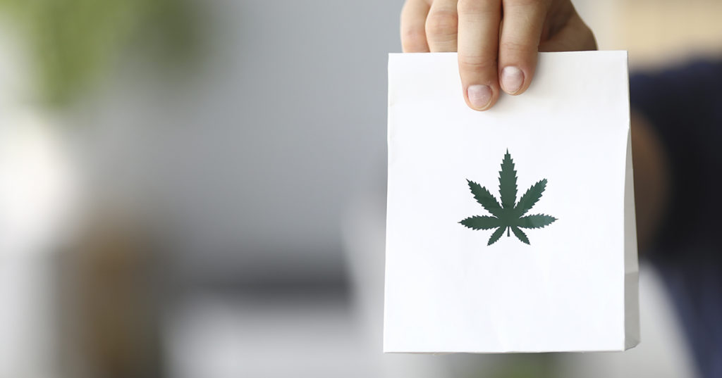 hand holding out a small white paper bag with a green cannabis leaf printed on the center