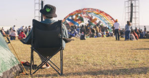 A person at an outdoor festival sitting on a foldable camp chair, wearing a hat with a visible cannabis leaf